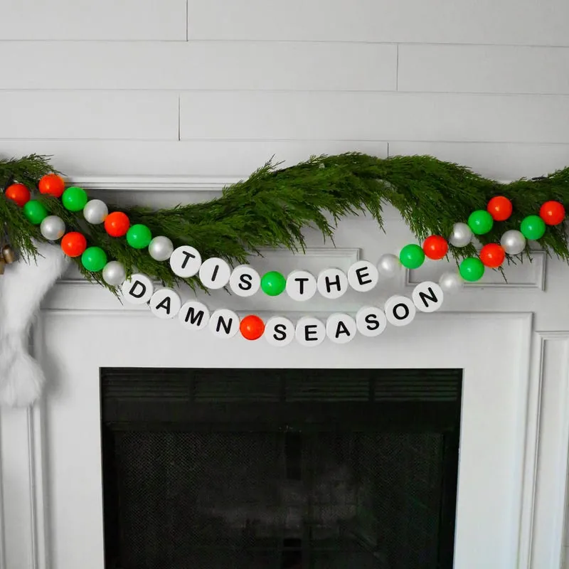 Bead garland spelling 'tis the damn season across a mantel